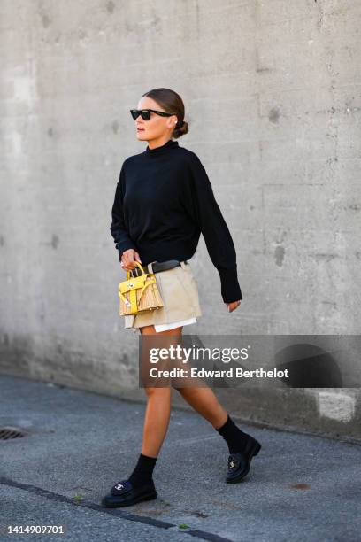 Guest wears black sunglasses, a black high neck pullover, a black shiny leather belt, a beige pleated / ripped short skirt from Miu Miu, a beige...