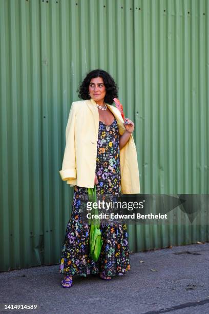 Guest wears white pearls double necklace, a pale yellow oversized blazer jacket, a black with multicolored flower print pattern V-neck long dress, a...