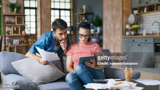 タブレットと電卓を使用して自宅で請求書を計算するカップル。ソファに座って財政を計算しながらコンピュータで働いている若いカップル。自宅で妻を持つ若い男性は、文書で彼らの財政� - カップル ストックフォトと画像