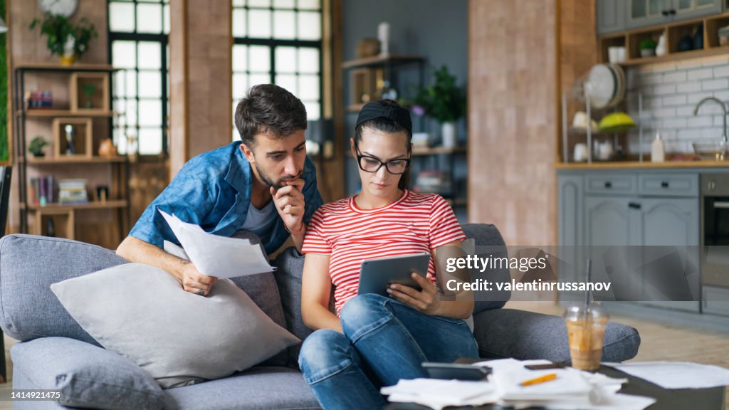 Couple calculating bills at home using tablet and calculator. Young couple working on computer while calculating finances sitting on couch. Young man with wife at home analyzing their finance with documents.