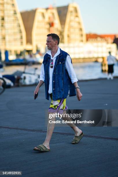 Guest wears a white shirt, a blue denim sleeveless / oversized gilet, purple and yellow print pattern shorts, khaki sandals, a silver watch , outside...