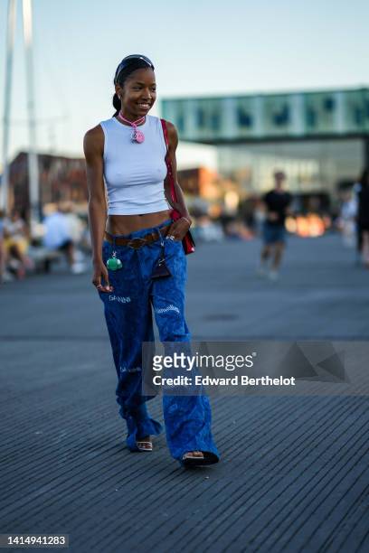 Guest wears black futurist sunglasses, a white cropped tank-top, a pink pendant necklace, a brown leather belt, a red shiny leather shoulder bag,...