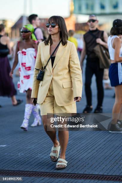 Guest wears sunglasses, a black shiny leather Lady D-light crossbody bag from Dior, a yellow blazer jacket, matching yellow shorts, white latte...