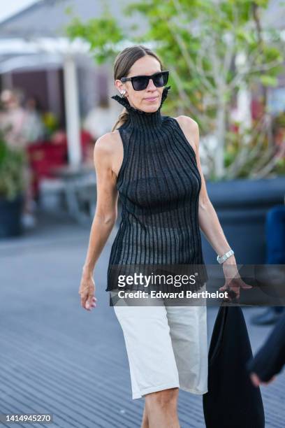 Guest wears black sunglasses, silver earrings, a black ribbed high neck / sleeveless t-shirt, a silver watch, white shorts, a black large handbag ,...