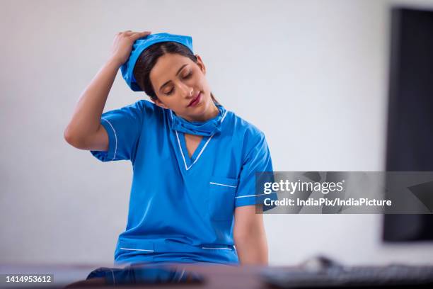 portrait of a tired female nurse removing surgical cap while sitting at computer desk - chirurgenkappe stock-fotos und bilder