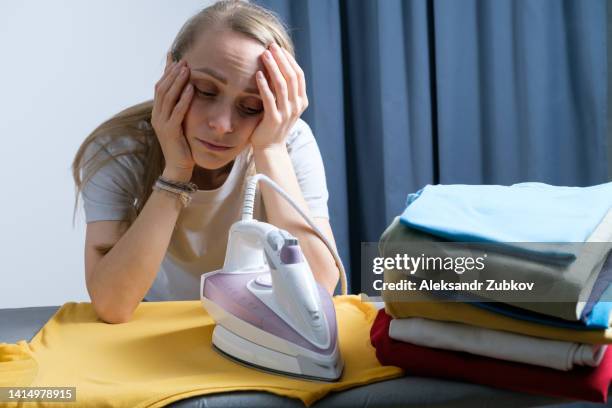 lots of bright, clean, multicolored ironed clothes stacked on the ironing board. a sad, exhausted, tired woman, housewife, cleaner or housekeeper is ironing a t-shirt. the concept of homework, lack of time for household chores. women's duties, home life. - strijkijzer stockfoto's en -beelden