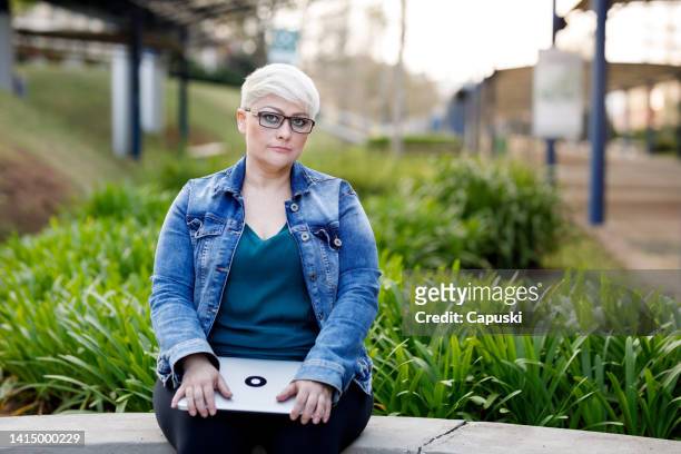 femme avec une coupe pixie et des lunettes à monture épaisse tenant un ordinateur portable assis sur le campus - coupe pixie photos et images de collection