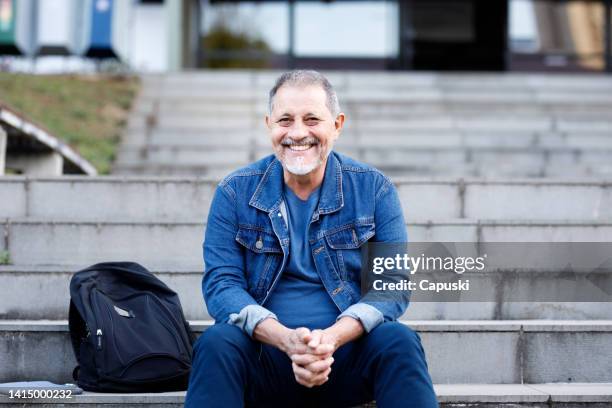 man with grey hair and beard smiling while sitting on the university stairs with his packpack - community college campus stock pictures, royalty-free photos & images