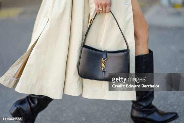 Guest wears a beige long buttoned trench coat, black shiny leather pointed / block heels knees boots / high boots, a black shiny leather with gold...