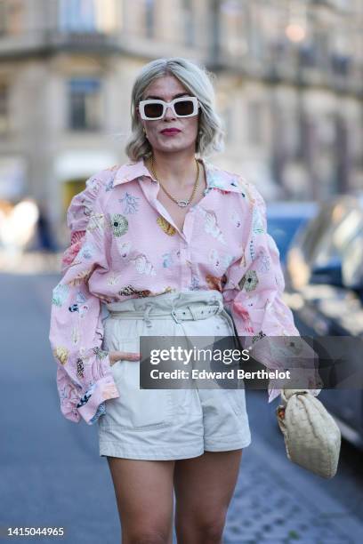 Guest wears pale pink square sunglasses, gold and pearl pendant earrings, a gold chain necklace, a pale pink with multicolored shell print pattern...