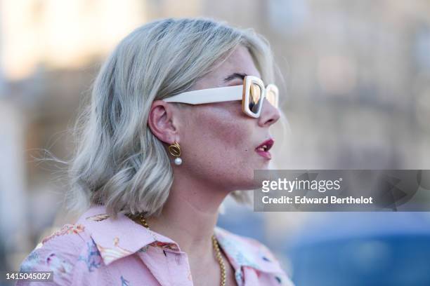 Guest wears pale pink square sunglasses, gold and pearl pendant earrings, a gold chain necklace, a pale pink with multicolored shell print pattern...