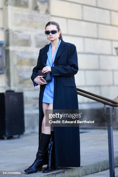 Guest wears black sunglasses, gold earrings, a blue cropped blazer jacket, matching blue suit short skirt, a black oversized long coat, a black shiny...