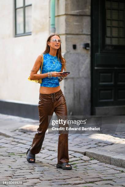 Guest wears black sunglasses, silver earrings, a blue tie and dye print pattern halter neck / cropped tank-top, a gold shiny leather shoulder bag,...