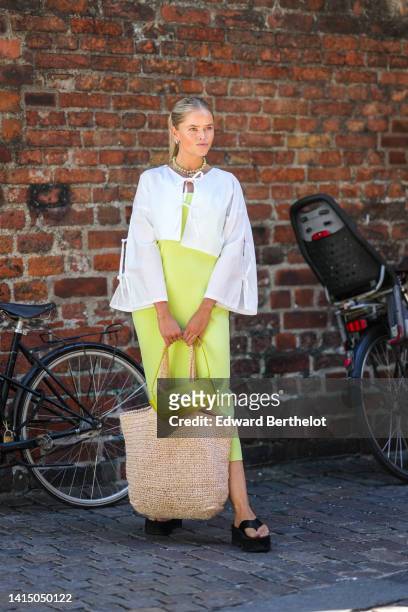 Guest wears silver earrings, a white oversized shirt, a beige with black embroidered logo oversized shoulder bag from Rotate Sunday, a pale green...