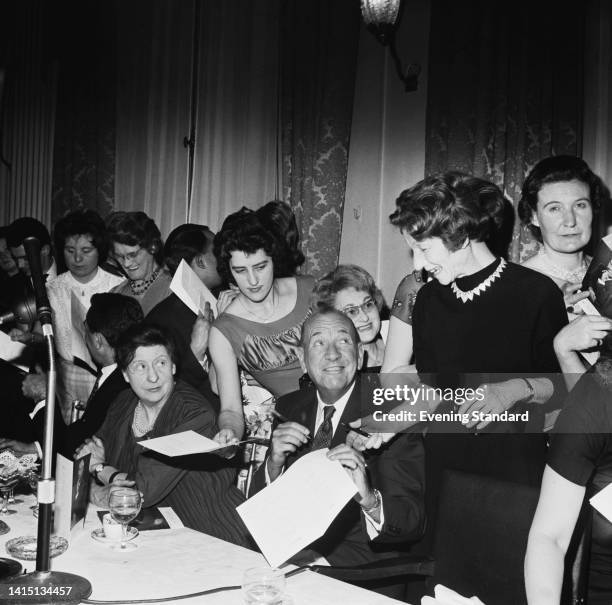 British actor, playwright and songwriter Noel Coward holding a sheet of paper as he is handed a pen, surrounded by women, United Kingdom, 21st May...