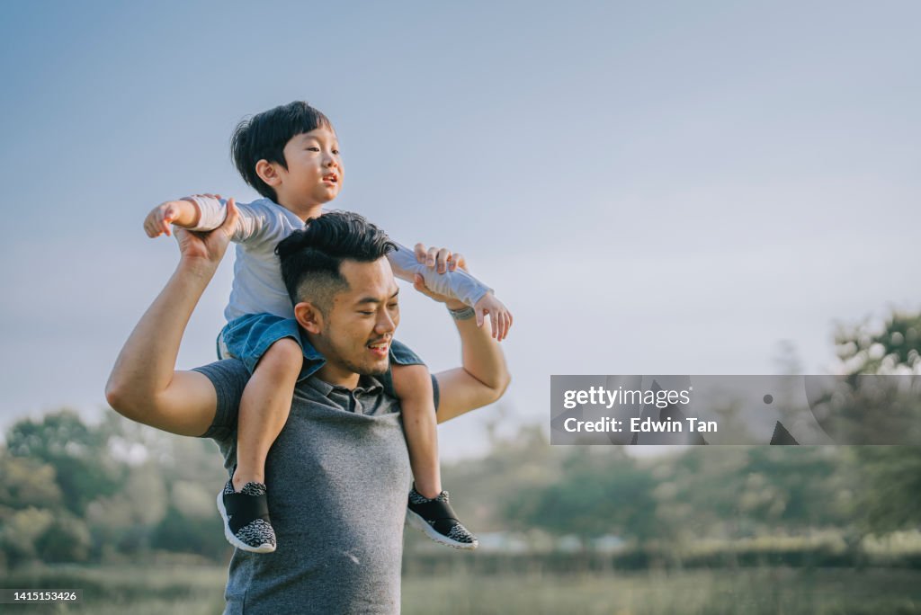 Asian chinese father carrying his son on shoulder at public park enjoying bonding time together during weekend leisure time