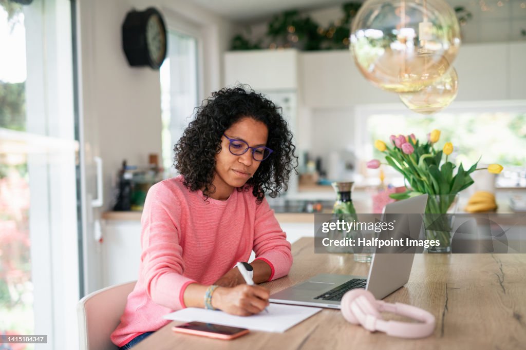 Multiracial woman having home office in kitchen, writing notes.