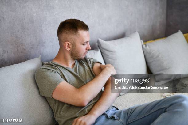 male picking at scab with fingers while sitting on couch - huidaandoening stockfoto's en -beelden