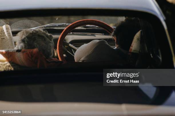 Man sleeps in the driving seat of a parked car, while a woman reads a newspaper in the passenger seat beside him, England, circa 1980.