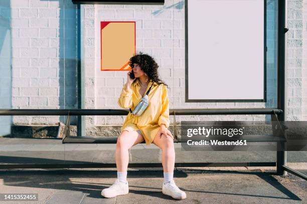 woman talking on smart phone sitting at bus stop - bushaltestelle stock-fotos und bilder