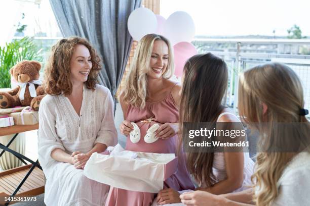 happy pregnant woman holding baby booties sitting by friends on sofa - babyshower stock-fotos und bilder