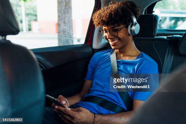 smiling young man wearing wireless headphones texting through smart phone - elektro auto stock-fotos und bilder