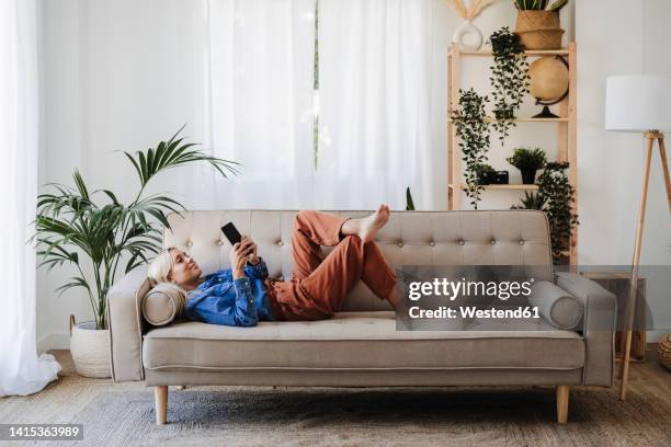 young woman using mobile phone lying on sofa at home - women lying imagens e fotografias de stock