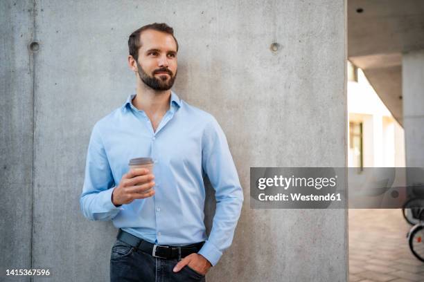 young businessman with coffee cup standing in front of wall - hands in pockets stock pictures, royalty-free photos & images