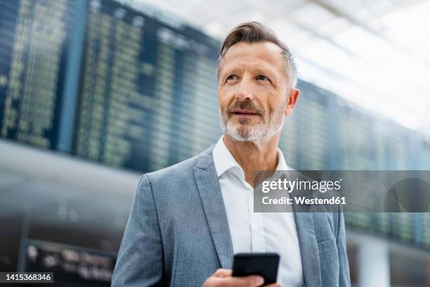 contemplative mature businessman holding smart phone - departure board front on fotografías e imágenes de stock