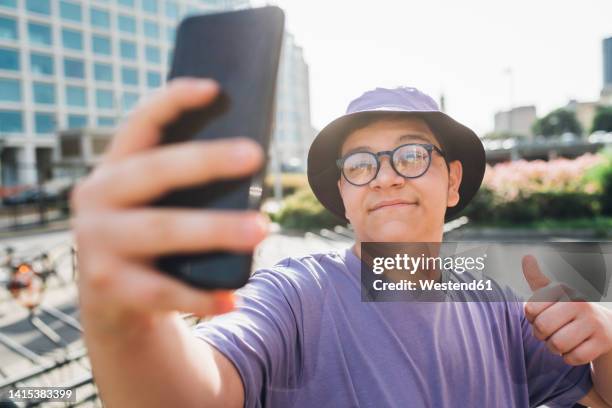 smiling teenage boy gesturing thumbs up and taking selfie on smart phone - bucket hat stock pictures, royalty-free photos & images