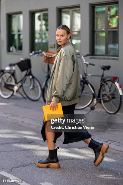 Mathilde Ravn is seen wearing yellow Copenhagen Studios bag, black brown Copenhagen Studios boots, black skirt with slit, olive bomber jacket on...