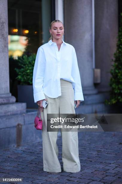 Guest wears gold earrings, a white oversized shirt, beige wide legs denim pants, gold rings, a pink shiny grained leather mini Timeless handbag from...