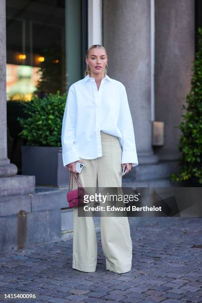Guest wears gold earrings, a white oversized shirt, beige wide legs denim pants, gold rings, a pink shiny grained leather mini Timeless handbag from...