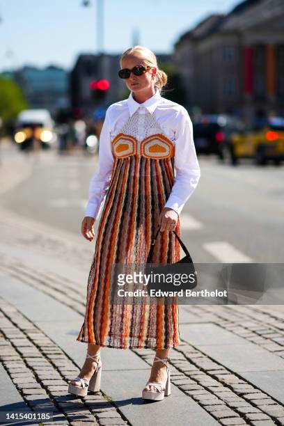 Guest wears brown sunglasses, a white fishnet / mesh yoke shirt, an orange and yellow striped print pattern shoulder-off long dress, a black shiny...