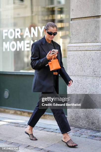 Guest wears gold earrings, a black blazer jacket, a black wool polo shirt, an orange shiny leather Kelly handbag from Hermes, silver diamonds rings,...