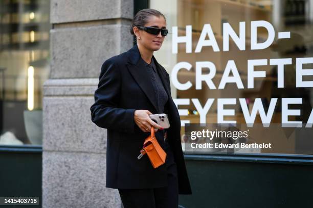 Guest wears gold earrings, a black blazer jacket, a black wool polo shirt, an orange shiny leather Kelly handbag from Hermes, silver diamonds rings,...