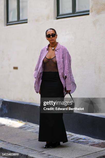 Guest wears black sunglasses, a black necklace with a silver heart pendant necklace, a burgundy and white striped print pattern oversized shirt, a...