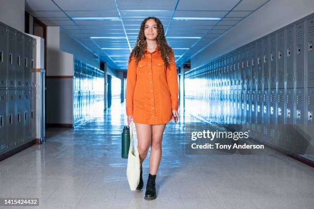 female student walking in high school corridor holding book bag and water bottle - shirt dress stock pictures, royalty-free photos & images