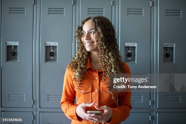 female high school student standing near lockers holding phone - hispanic teen girl stock pictures, royalty-free photos & images