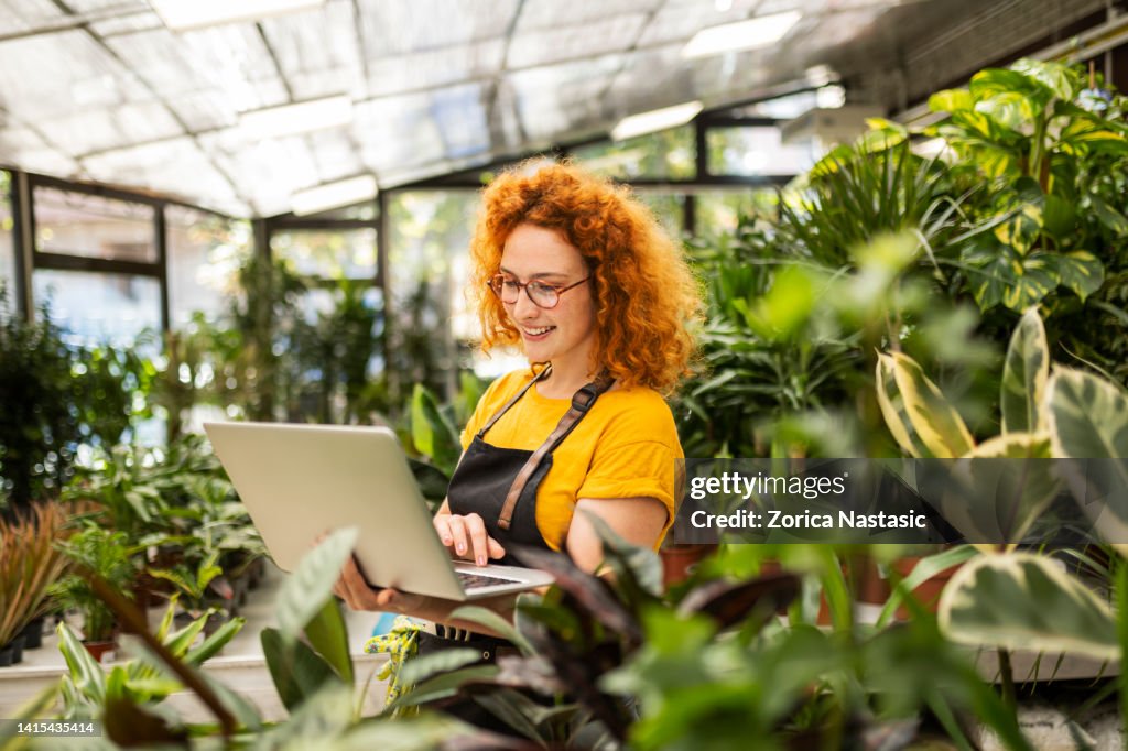 Young redhead woman working in a flower shop with potted houseplants holding laptop