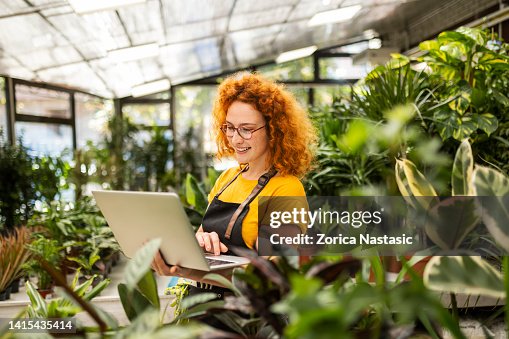 Young redhead woman working in a flower shop with potted houseplants holding laptop Young redhead woman working in a flower shop with potted houseplants holding laptop