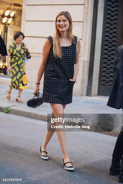 Eugenie Trochu of Vogue France wears a black dress with grommets and pockets, black and white low sung heels, and holds a black Alaia heart coeur...