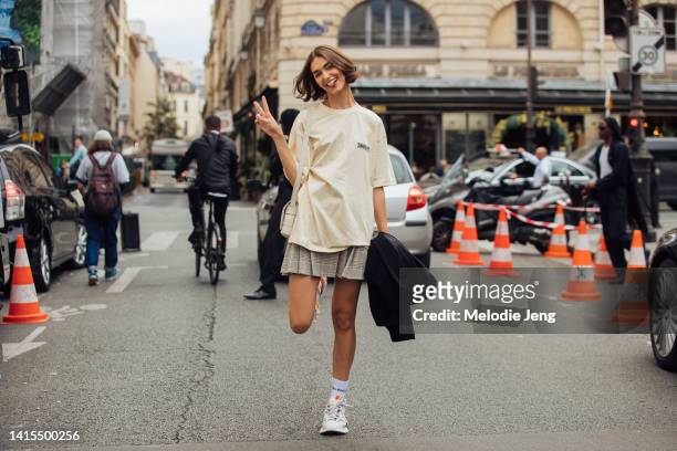 Model Rayssa Medeiros throws a peace sign and wears a beige Balenciaga political campaign large fit t-shirt, gray plaid pleated mini skirt, white...