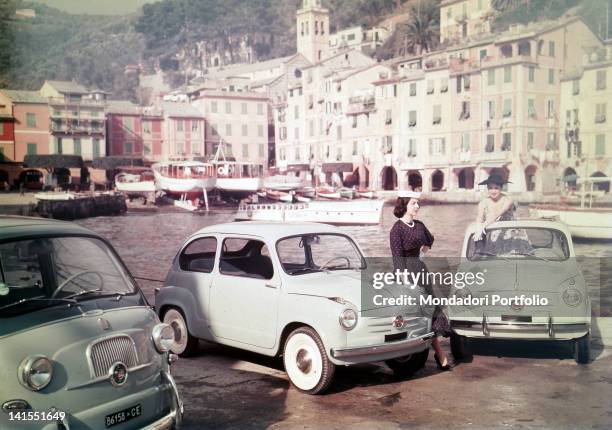 Two models posing in the port among three new Fiat 600 cars. Portofino, 1955