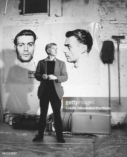 The American painter and director Andy Warhol posing in his studio in front of two portraits inspired by mug shots. New York, 1964