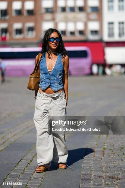 Guest wears black and blue sunglasses, a silver necklace, a white bra underwear, a blue denim faded V-neck / sleeveless / buttoned gilet, a brown...