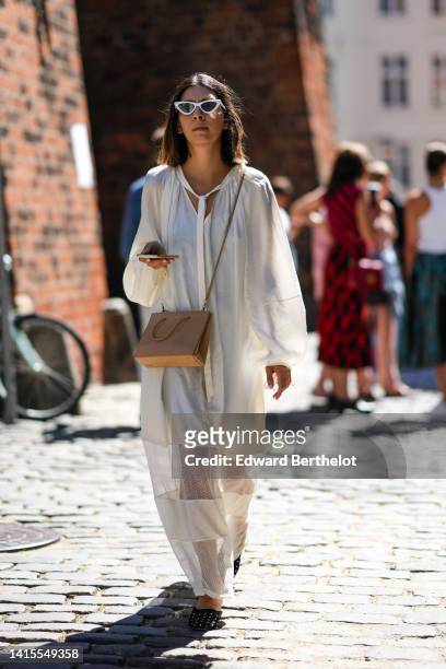 Guest wears white cat eyes sunglasses, a white oversized long sleeves / long dress with transparent large straps, a beige matte leather crossbody bag...