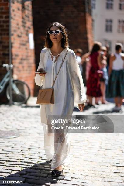 Guest wears white cat eyes sunglasses, a white oversized long sleeves / long dress with transparent large straps, a beige matte leather crossbody bag...