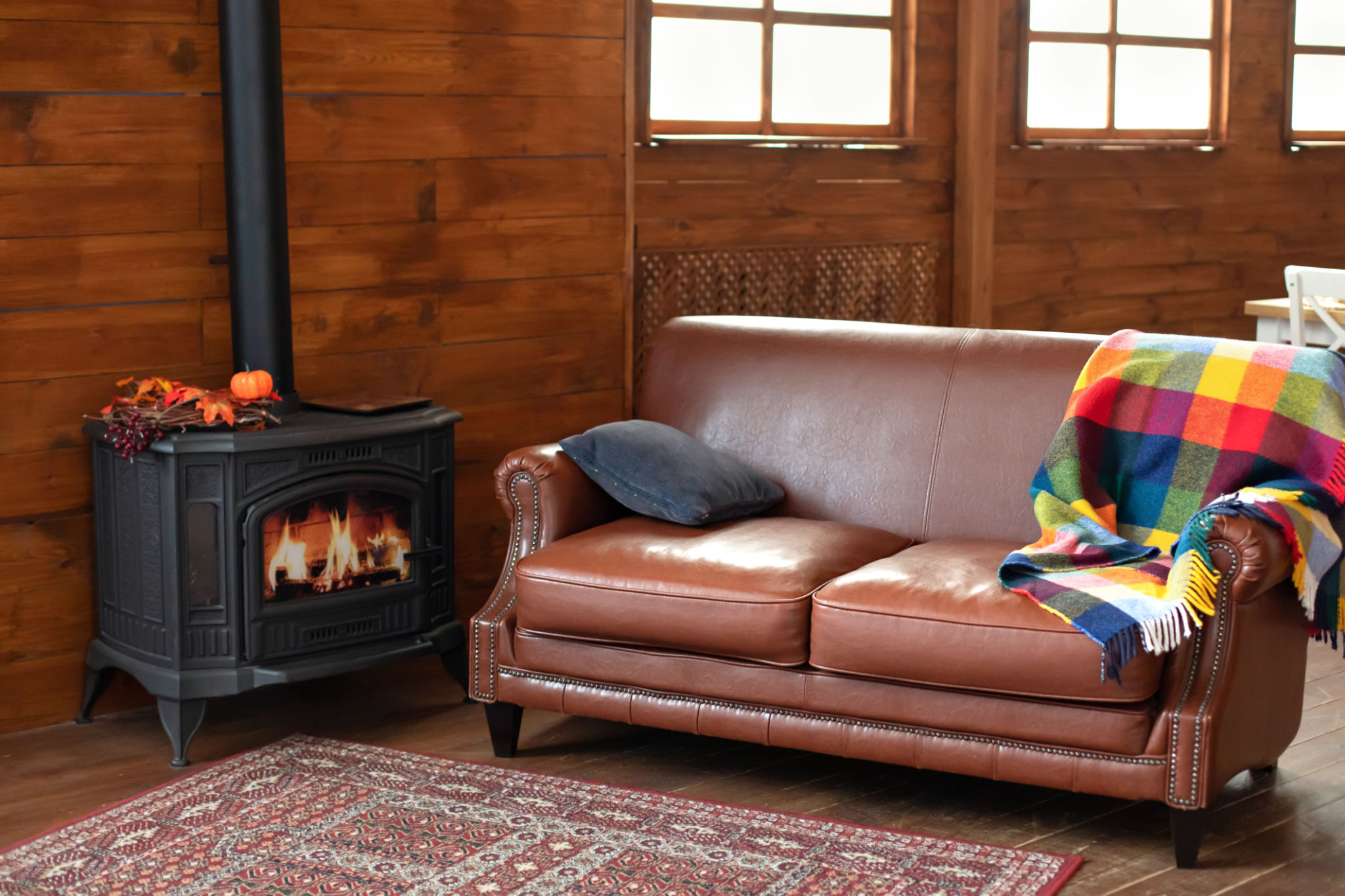 Interior cozy living room with wood burning stove inside, comfortable sofa, rustic wooden walls and carpet on floor. Black modern Cast iron wood stove at home. Fragment of interior of country house. Interior cozy living room with wood burning stove inside, comfortable sofa, rustic wooden walls and carpet on floor. Black modern Cast iron wood stove at home. Fragment of interior of country house.