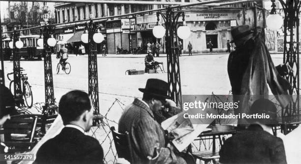 Austrian civilians sitting in an outdoor cafT protected by barbed wire, in the road a German soldier behind a machine gun.áVienna, February 1934